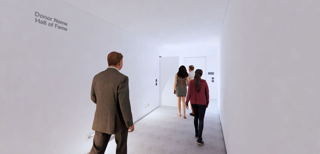 Five people walk down a bright, minimalistic hallway with white walls. The words "Donor Name Hall of Fame" are displayed on the left wall. The group is moving toward a door at the far end of the corridor.