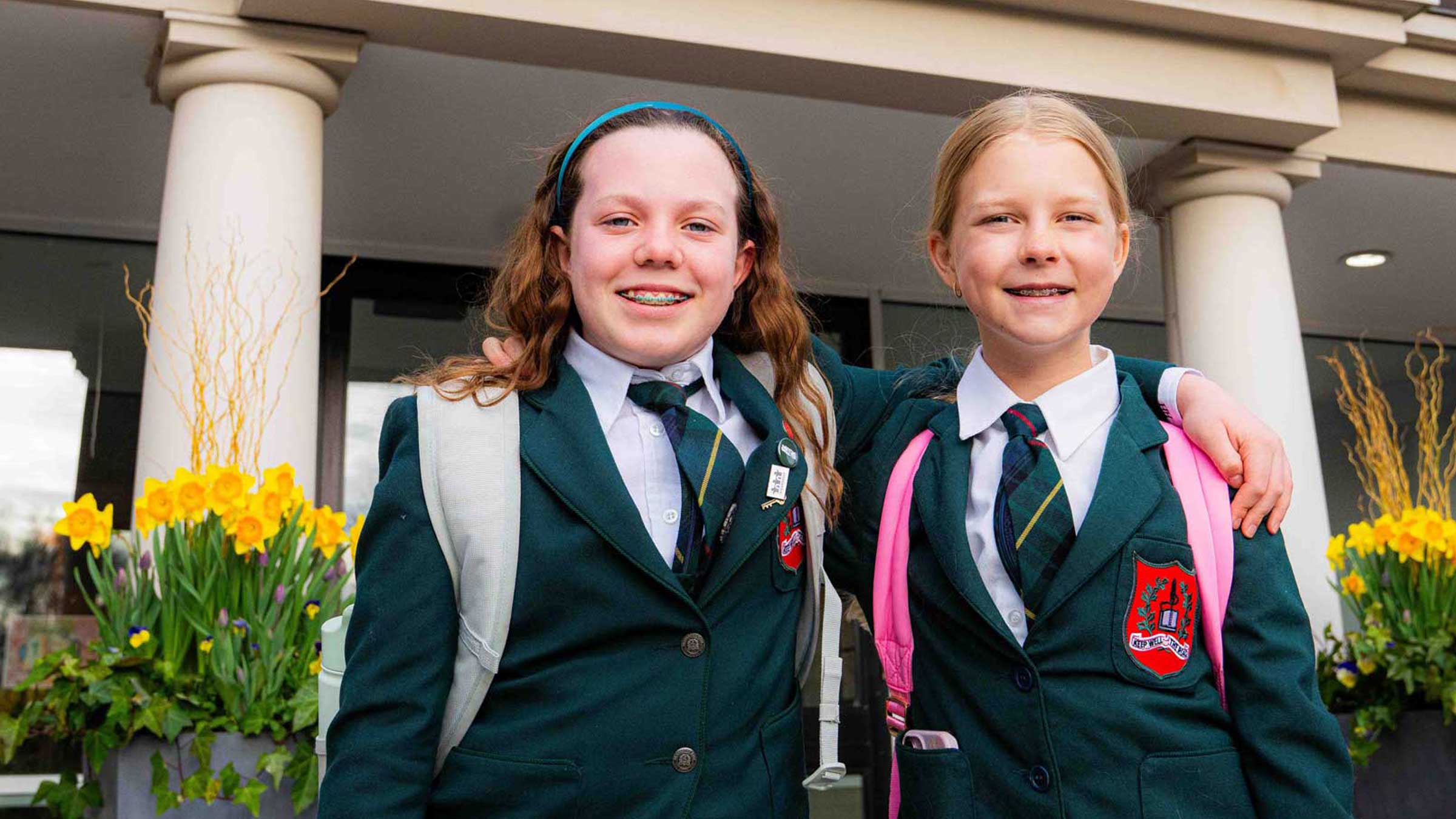 Two smiling schoolgirls in green uniforms with ties and backpacks stand together outside a building with columns and flower pots, arms around each other.