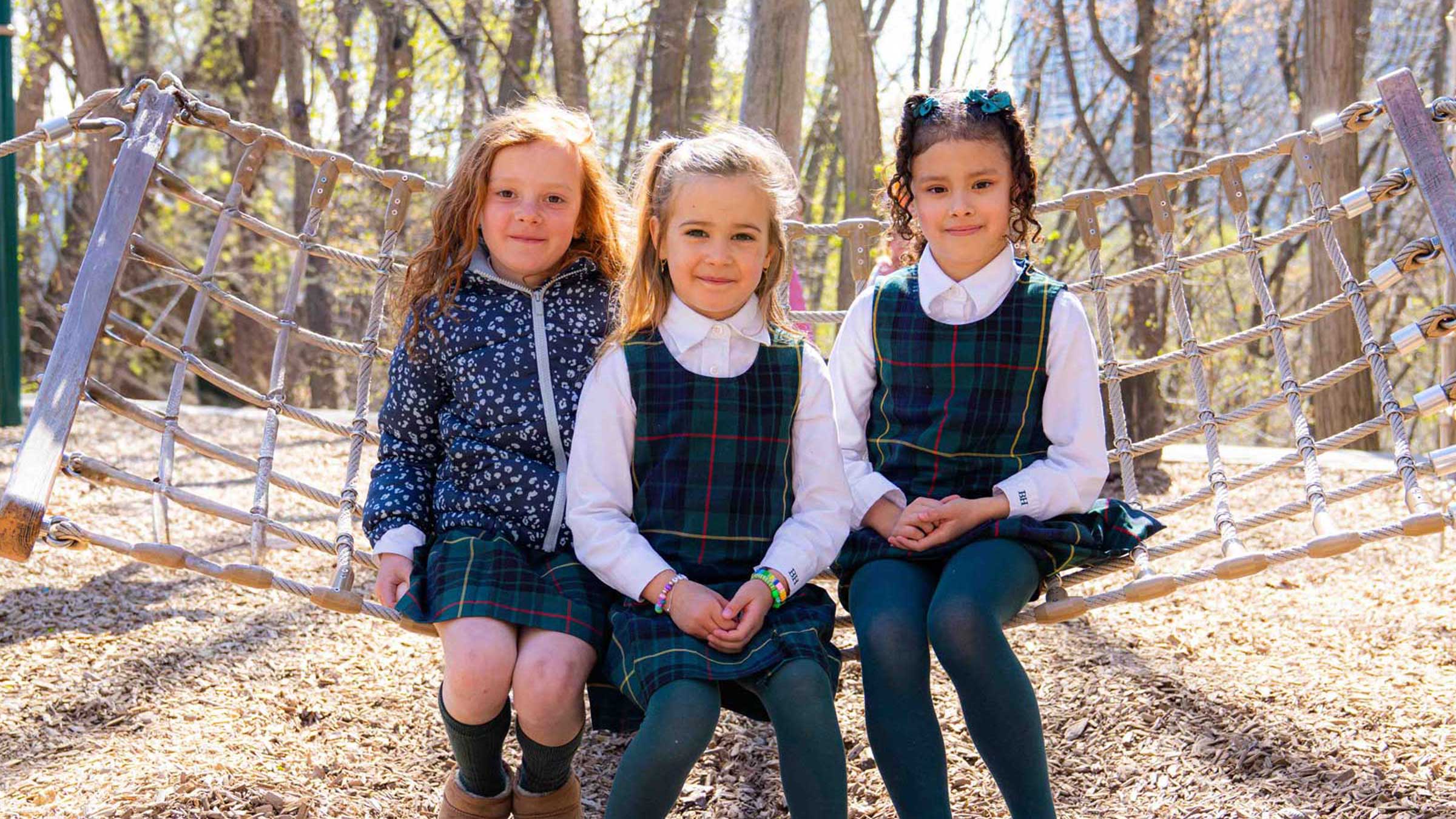 Three young girls sit together on a rope swing in a wooded area, smiling at the camera. Two wear matching plaid jumpers and collared shirts, while the third wears a blue patterned jacket over a dress. Sunlight filters through the trees.