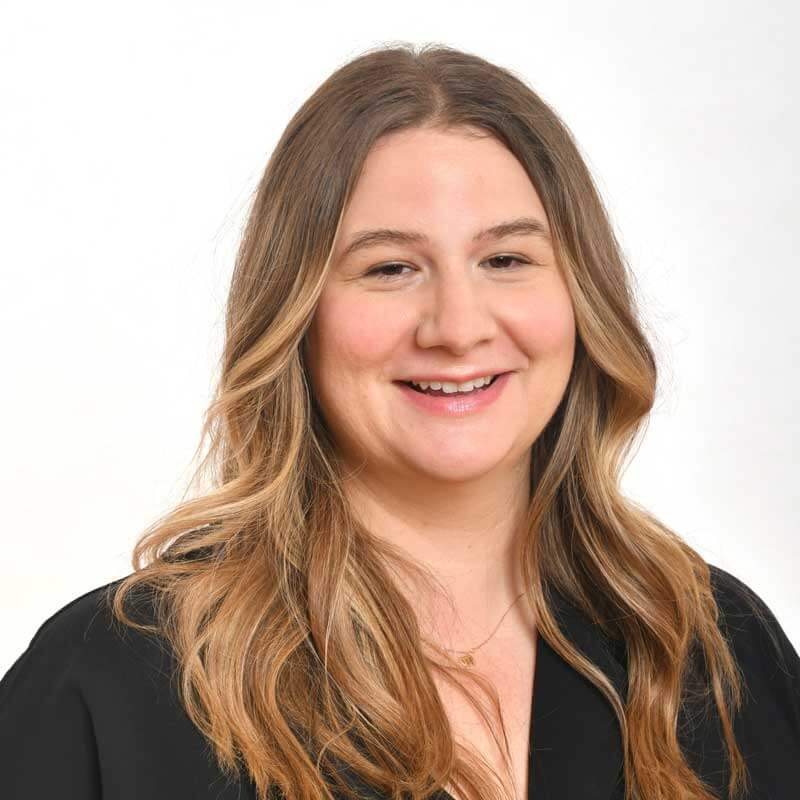 A woman with long, wavy light brown hair wearing a black top smiles at the camera against a plain white background.