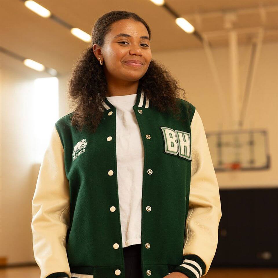 A young woman wearing a green and beige varsity jacket stands confidently in a gymnasium, with a basketball hoop visible in the background. She is smiling gently and looking at the camera.