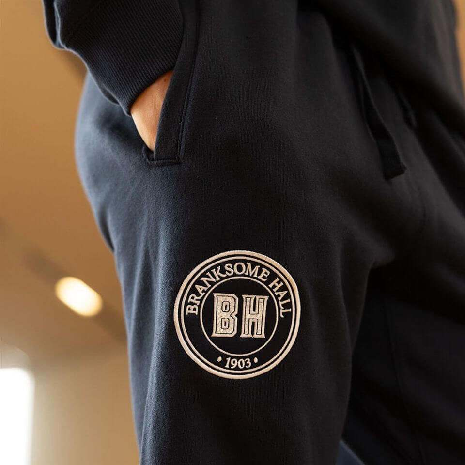 Close-up of a person wearing black sweatpants with one hand in the pocket. The sweatpants feature a round embroidered "Branksome Hall BH 1903" logo patch on the thigh.