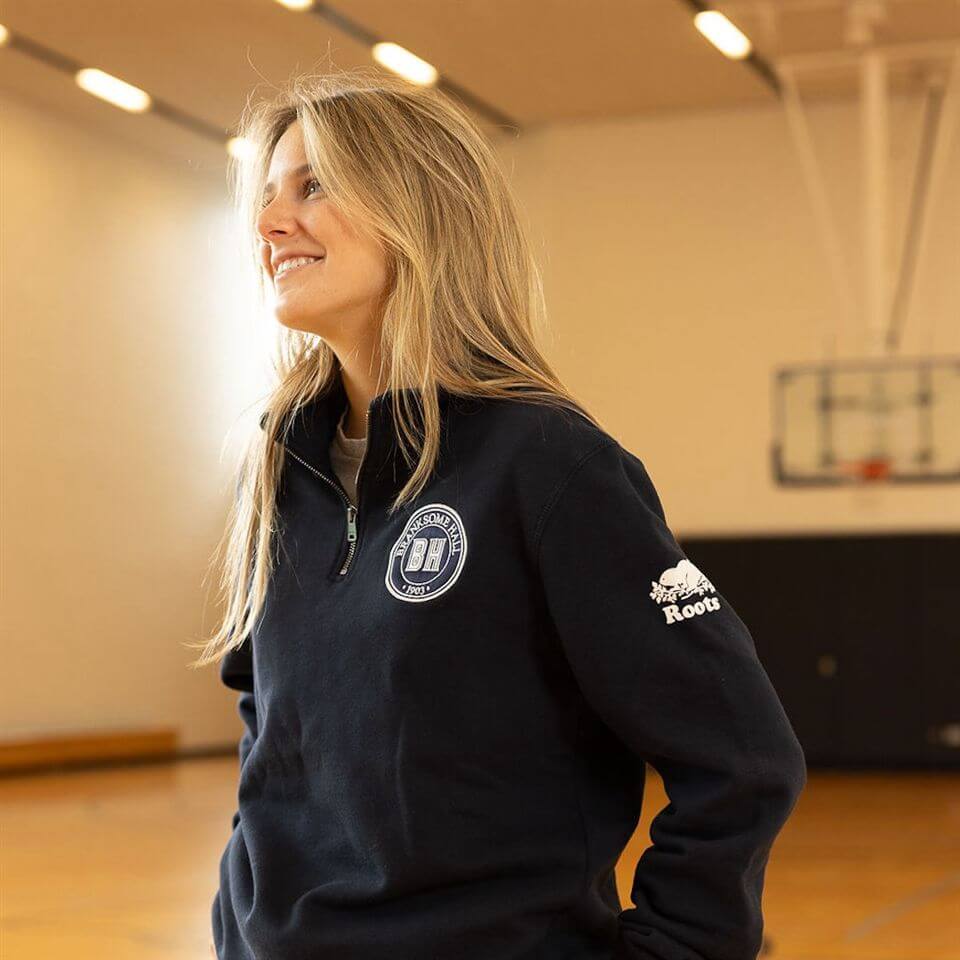 A young woman with long blonde hair smiles while standing in a gymnasium, wearing a navy pullover with a crest on the chest and "Roots" on the sleeve. A basketball hoop and wooden floor are visible in the background.