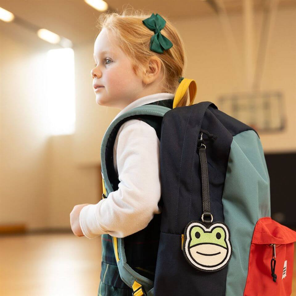 A young girl with a green bow in her hair wears a backpack with a frog face keychain, standing in a gymnasium with blurred basketball hoops in the background.