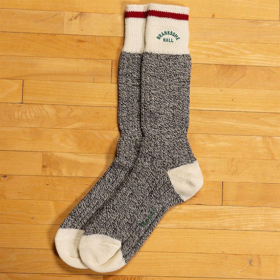 A pair of long, gray and white marled socks with cream-colored toes, heels, and cuffs, and a red stripe near the top. The words "DRAM & SMOKE HALL" are embroidered in green on the cuffs. The socks are laid on a wooden floor.