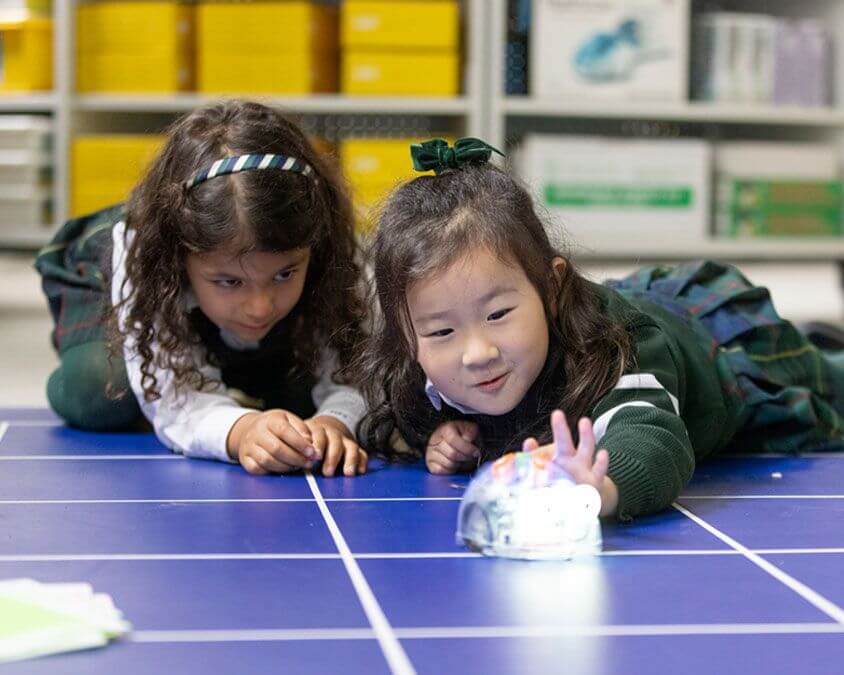 Two young girls in school uniforms lie on a blue grid floor, smiling and watching a small robotic toy move, with shelves and yellow bins in the background.