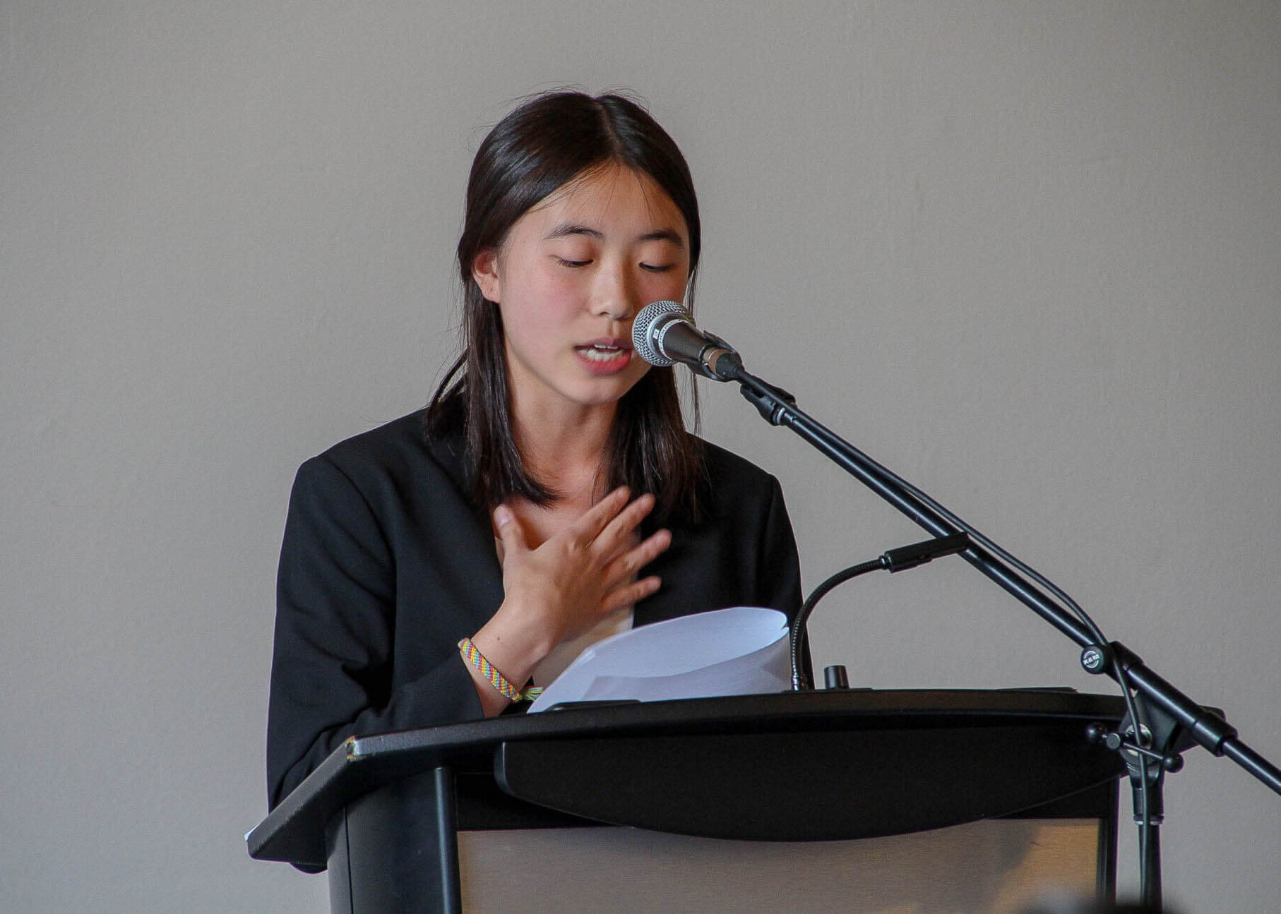 A young woman wearing a black blazer speaks into a microphone at a podium, holding papers in one hand and gesturing with her other hand on her chest against a plain background.