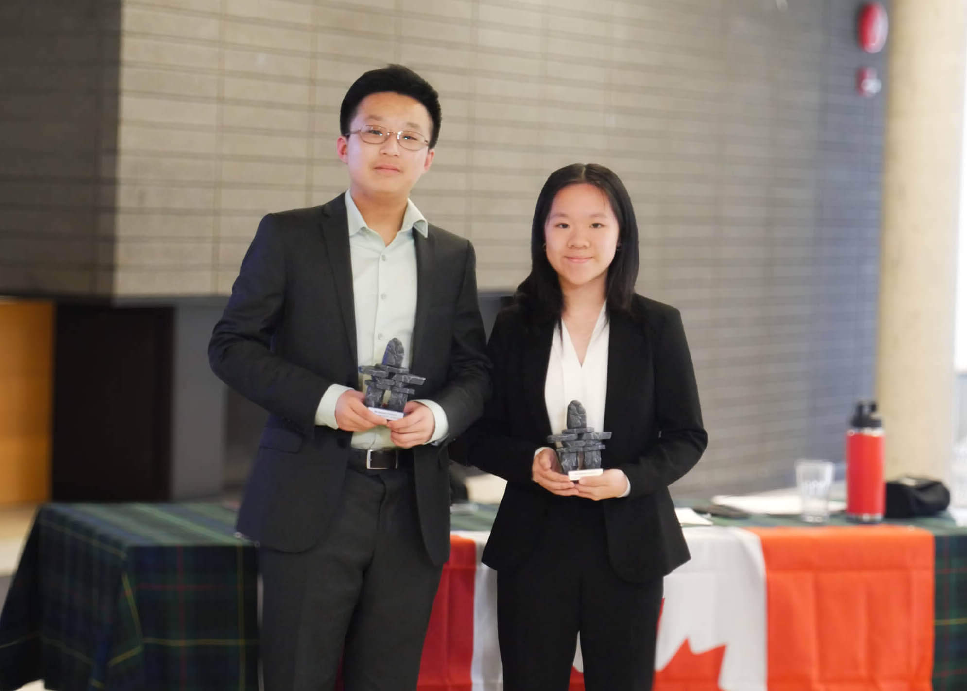 Two people in business attire stand indoors holding small trophies. Behind them is a table covered with a Canadian flag, a water bottle, and a checkered cloth, and a brick wall is in the background.