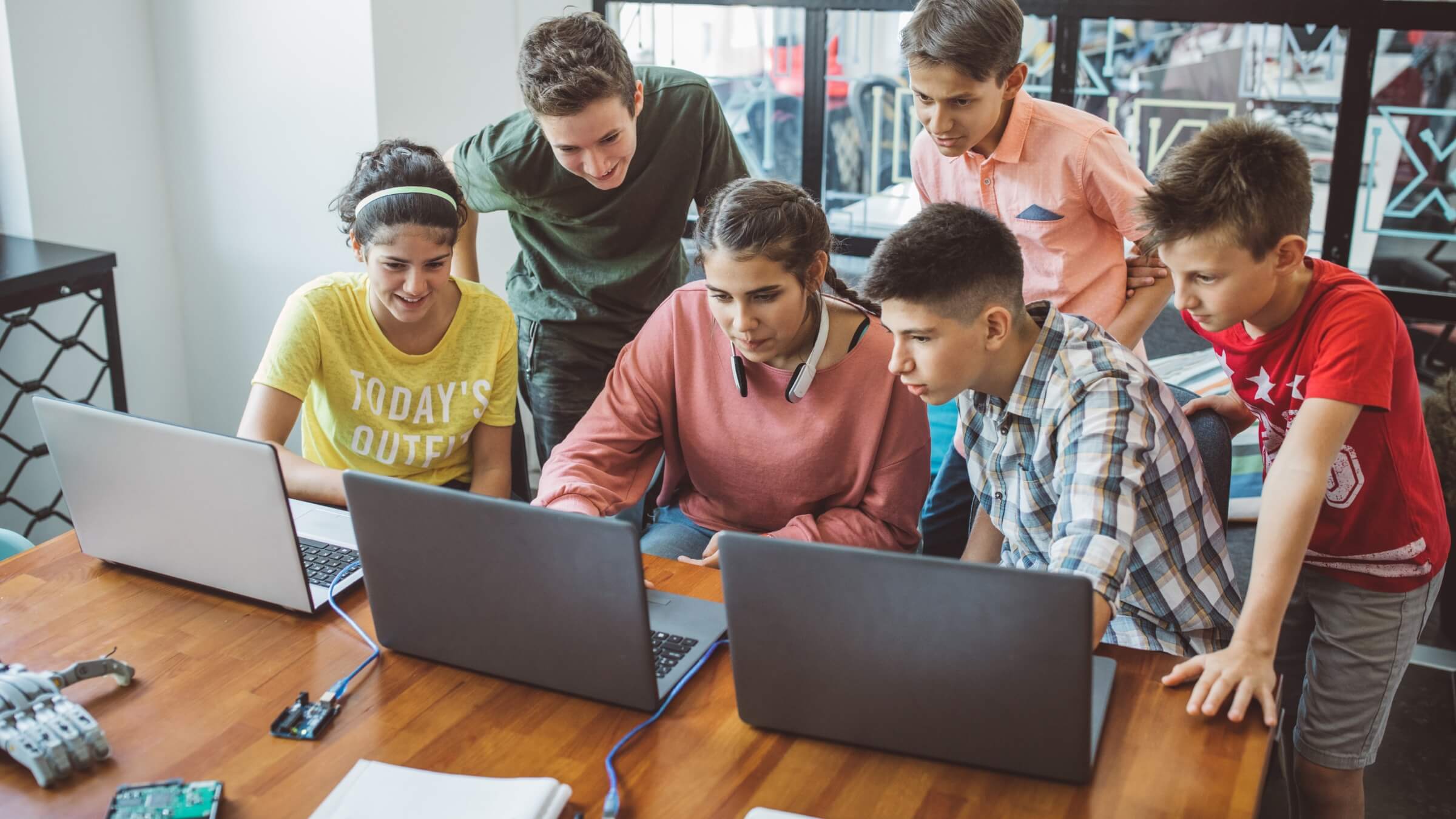 Six teenagers gathered around a table with three laptops, collaborating and looking engaged, suggesting teamwork on a technology or school project in a bright, modern room.