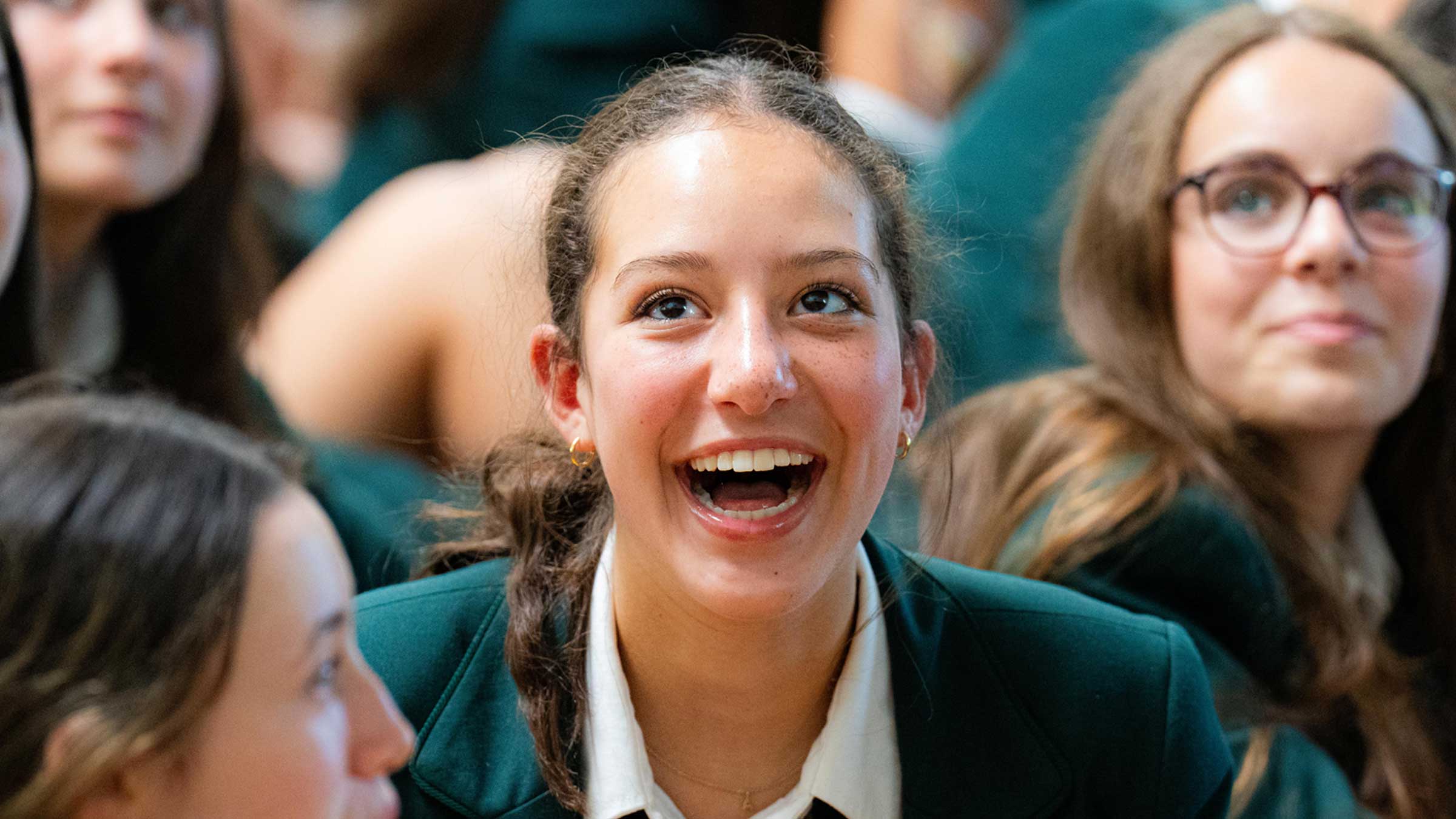 A teenage girl in a green school uniform smiles widely, surrounded by other students in similar attire. The atmosphere appears lively and cheerful.