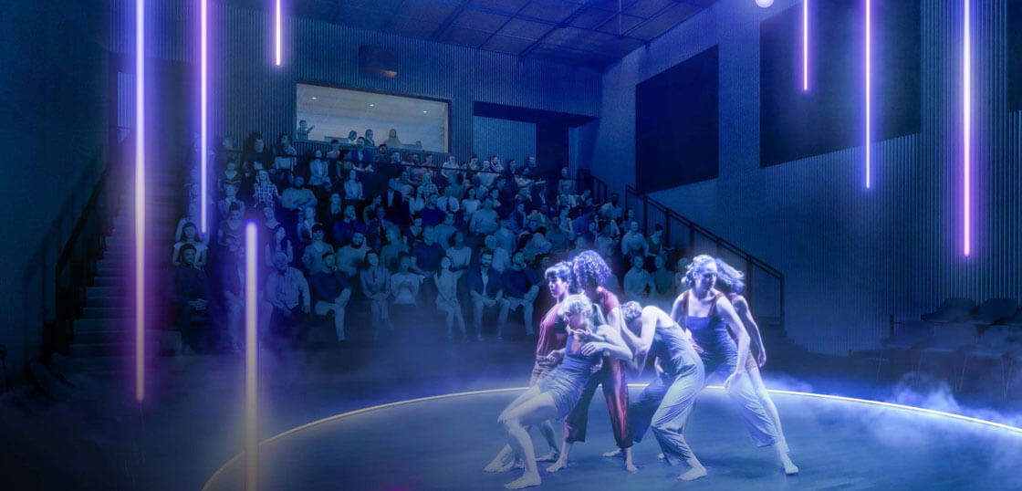 Four dancers perform on a circular, smoke-filled stage with glowing vertical lights, while a seated audience watches in a dimly lit modern theater.