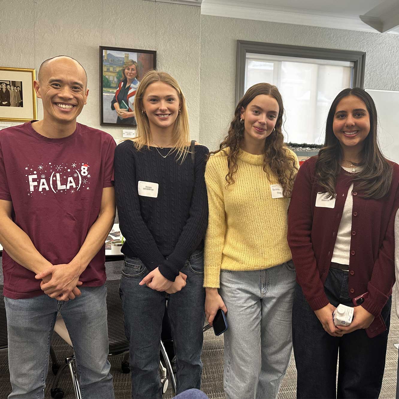 Four people stand indoors, smiling at the camera. The man on the left wears a maroon shirt, while the three women beside him wear sweaters in black, yellow, and burgundy. There are name tags on their chests and office decor in the background.