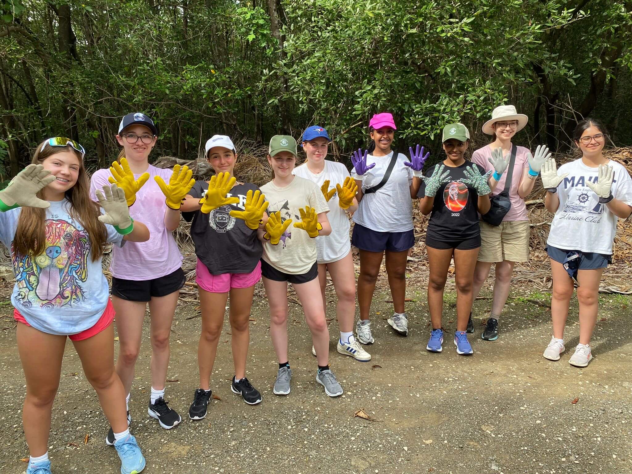 A group of nine smiling people stand outdoors on a dirt path, wearing colorful work gloves and casual clothes, with trees and greenery in the background. They hold up their gloved hands toward the camera.