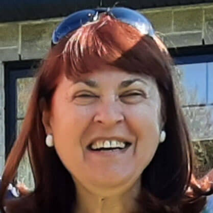 A woman with red hair, wearing white earrings and sunglasses on her head, smiles broadly outdoors in front of a building with large windows.