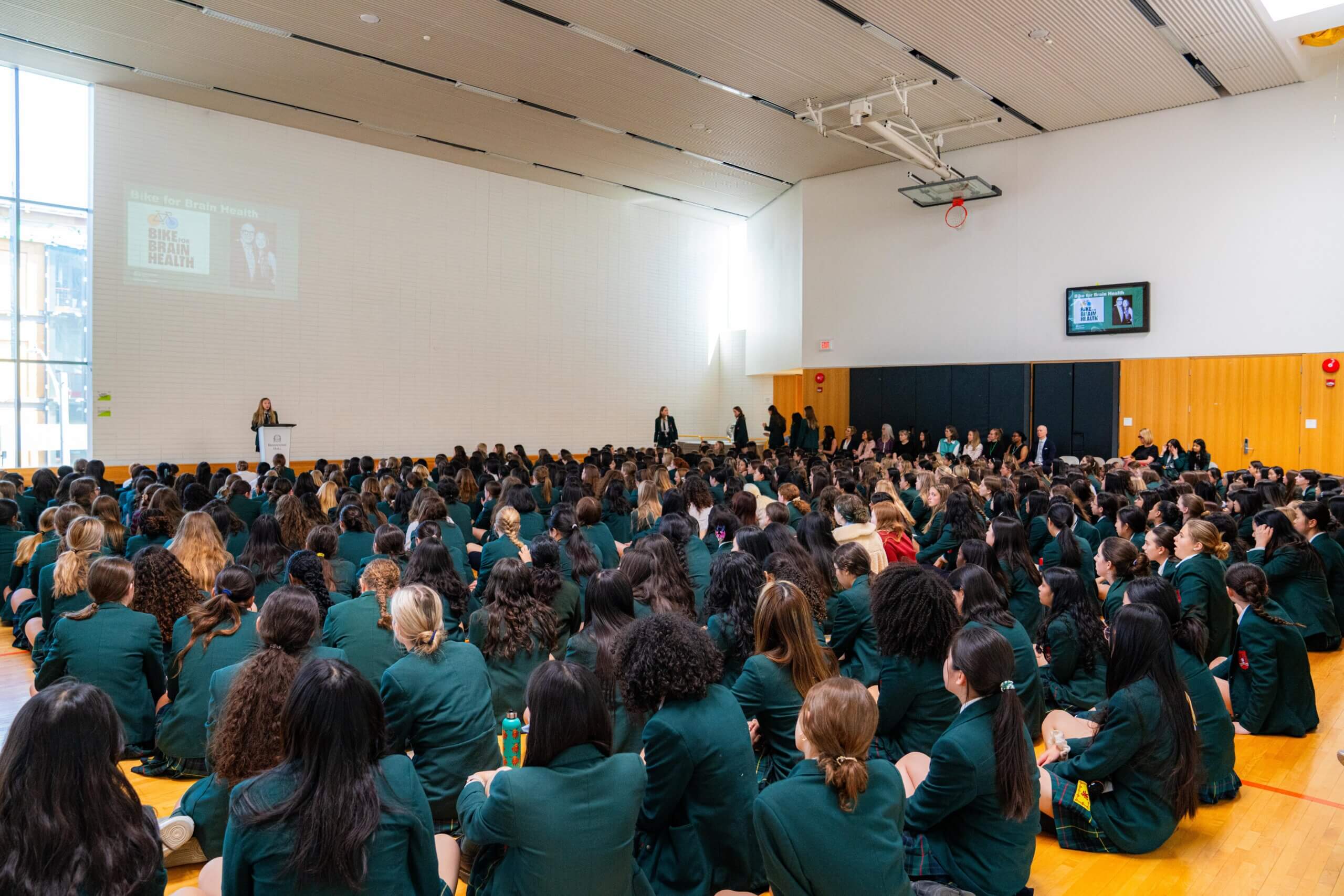 A large group of students in green uniforms sit on the floor of a gymnasium, facing a podium where a speaker presents near a projected image on the wall. The space is bright with wood paneling and high ceilings.