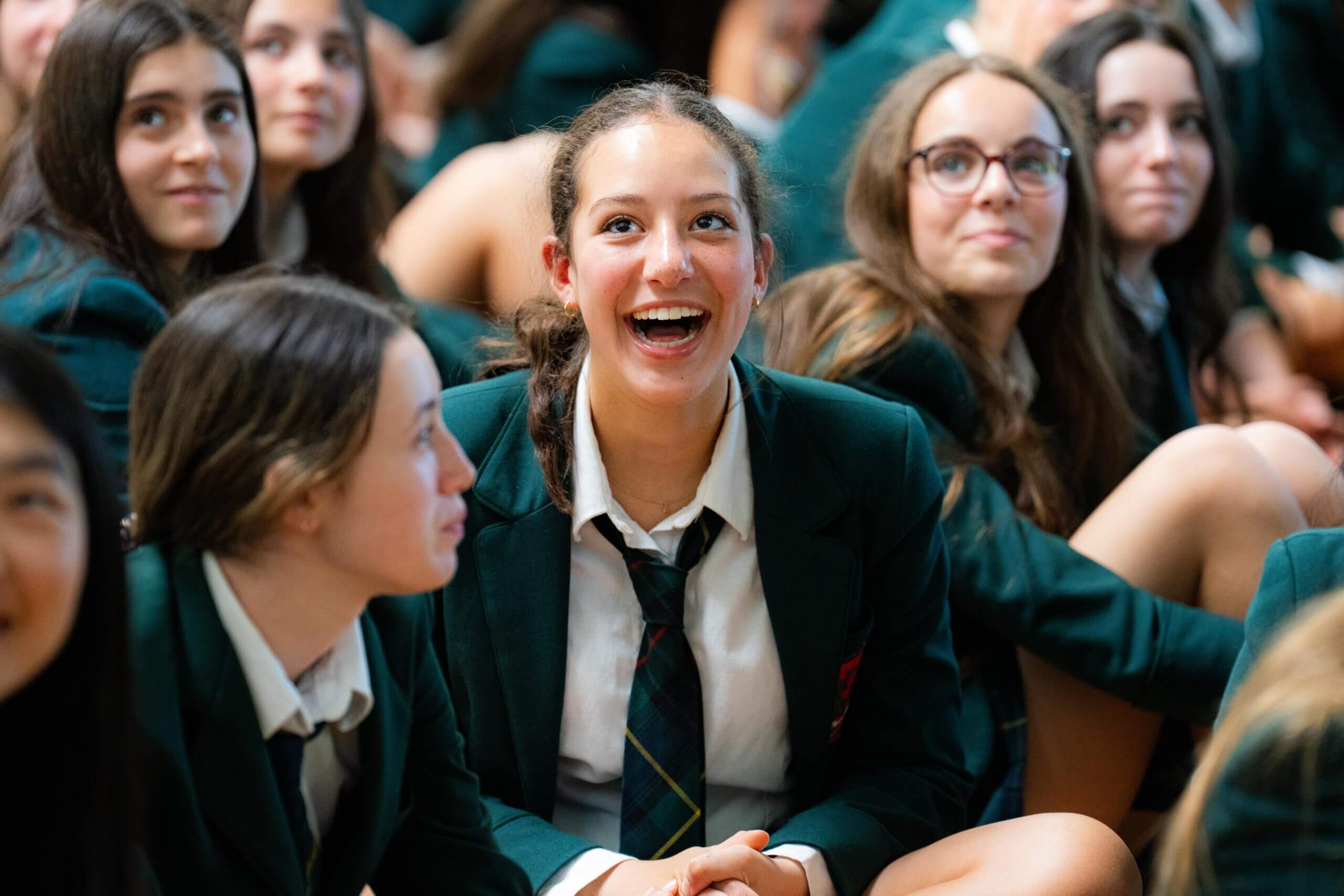 A group of students in green blazers and ties sit closely together, smiling and looking upward. The central girl is laughing, creating a joyful and lively atmosphere.