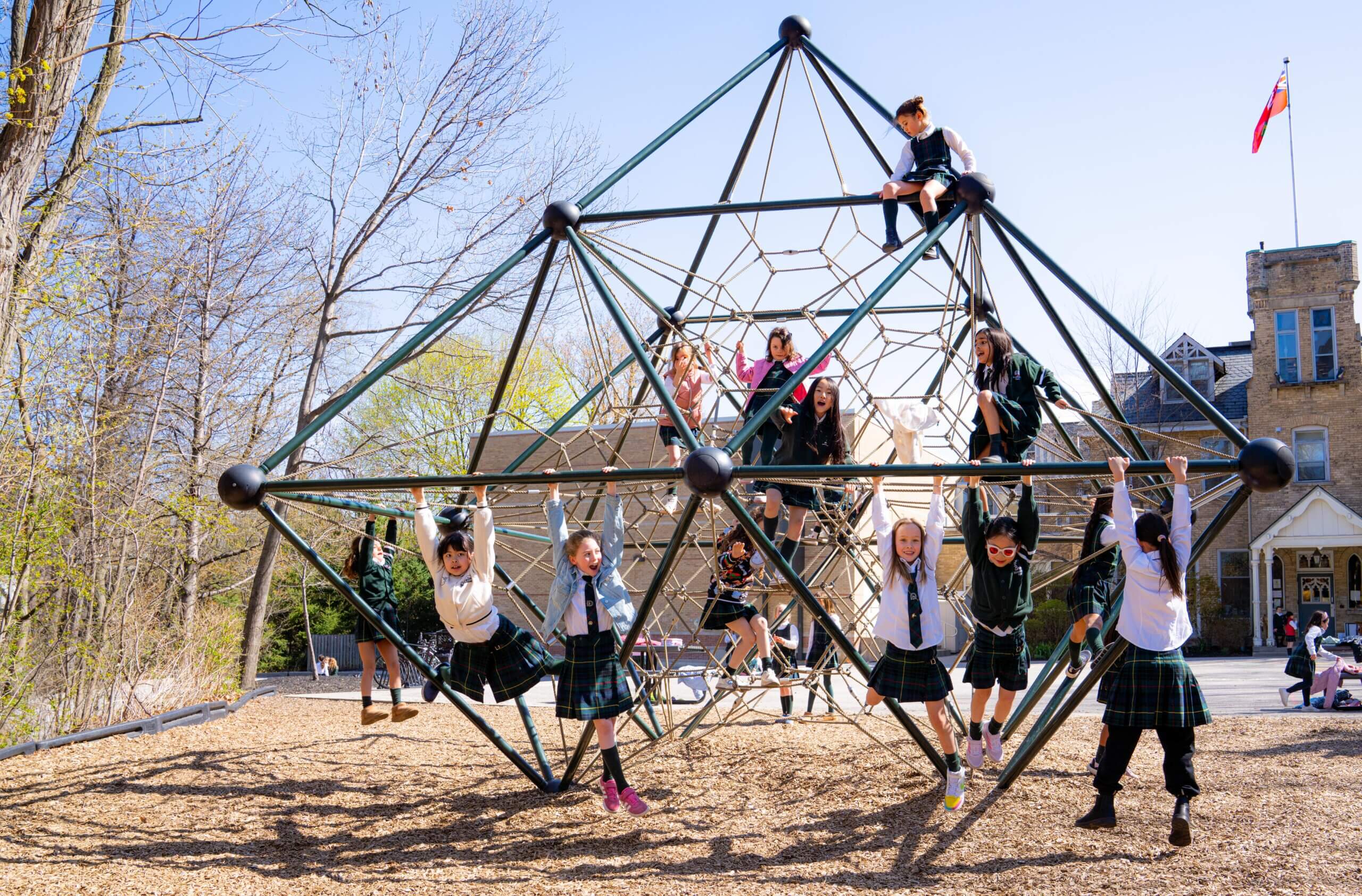 Children in school uniforms climb and play on a large geometric jungle gym outdoors on a sunny day, with trees and a school building in the background.