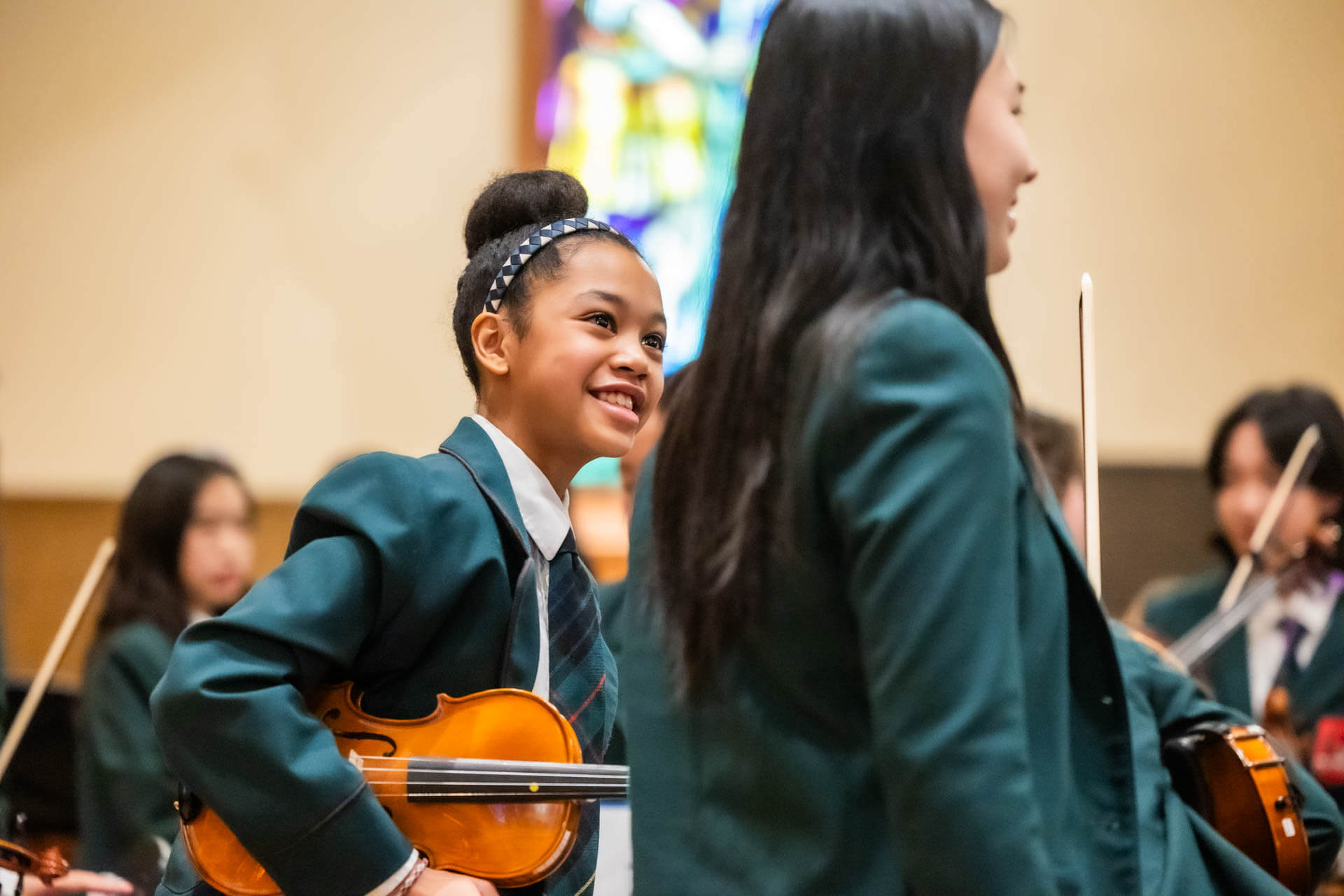 A smiling girl in a green school uniform holds a violin among other students in similar attire, with a blurred indoor background and colorful stained glass.
