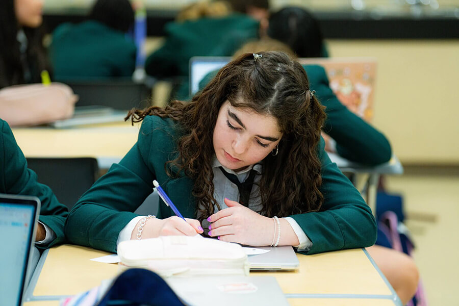 A student with long curly hair, wearing a green school blazer, sits at a desk and writes in a notebook. Other students are visible in the background, also working at their desks.