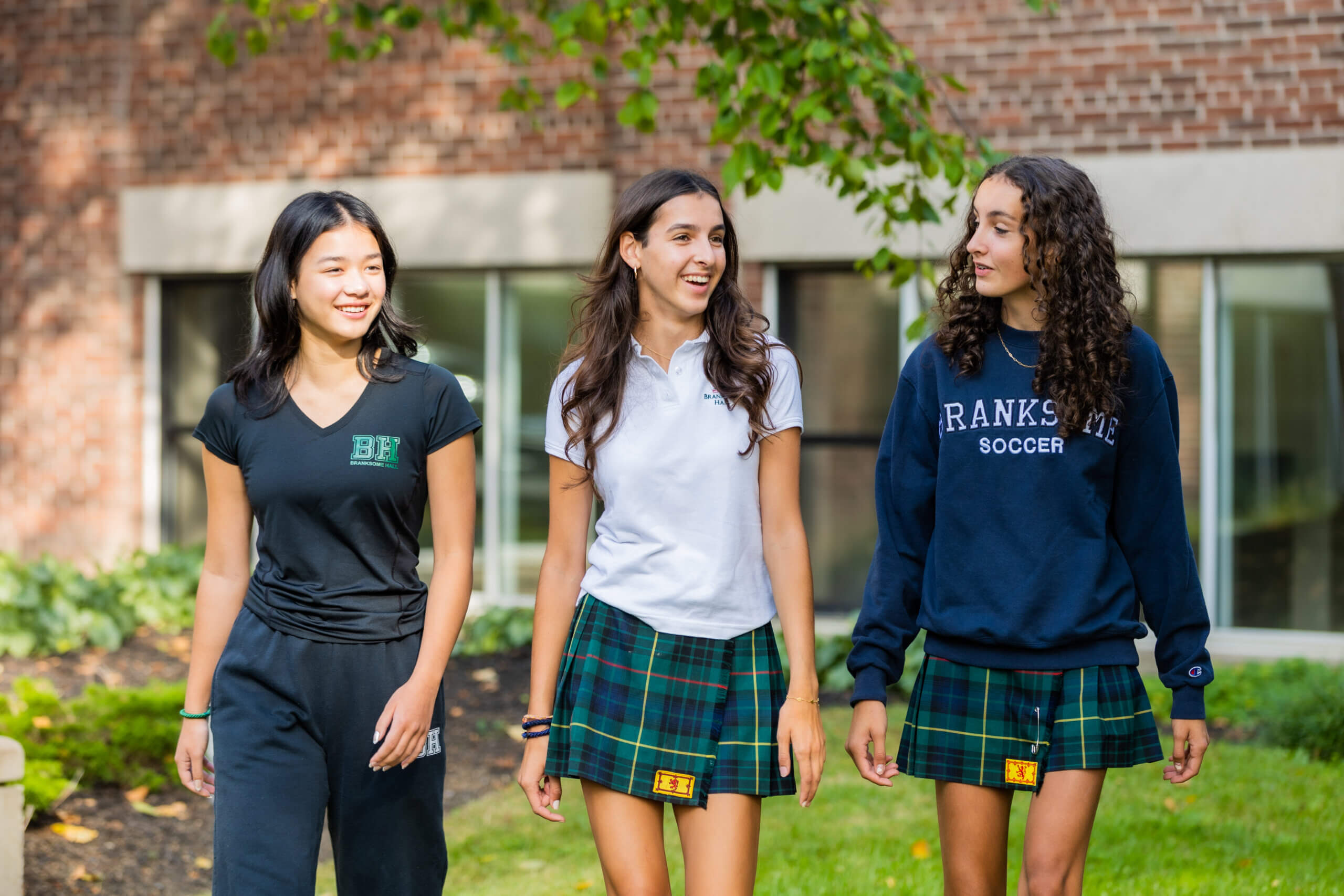 Three teenage girls in school uniforms walk outside on a sunny day. Two wear plaid skirts and one wears black pants. They are smiling and talking, with a brick building and greenery in the background.