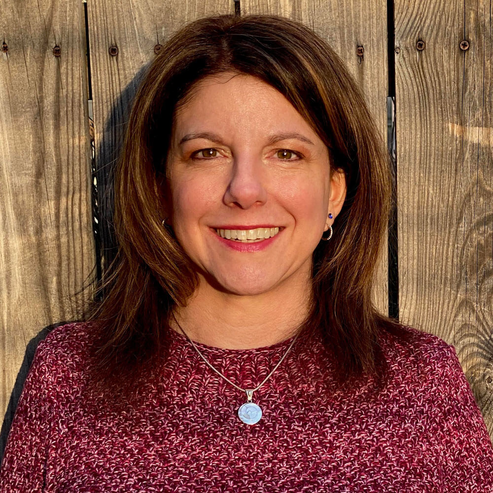A woman with shoulder-length brown hair, wearing a maroon patterned sweater and a silver necklace, smiles in front of a wooden fence in natural sunlight.