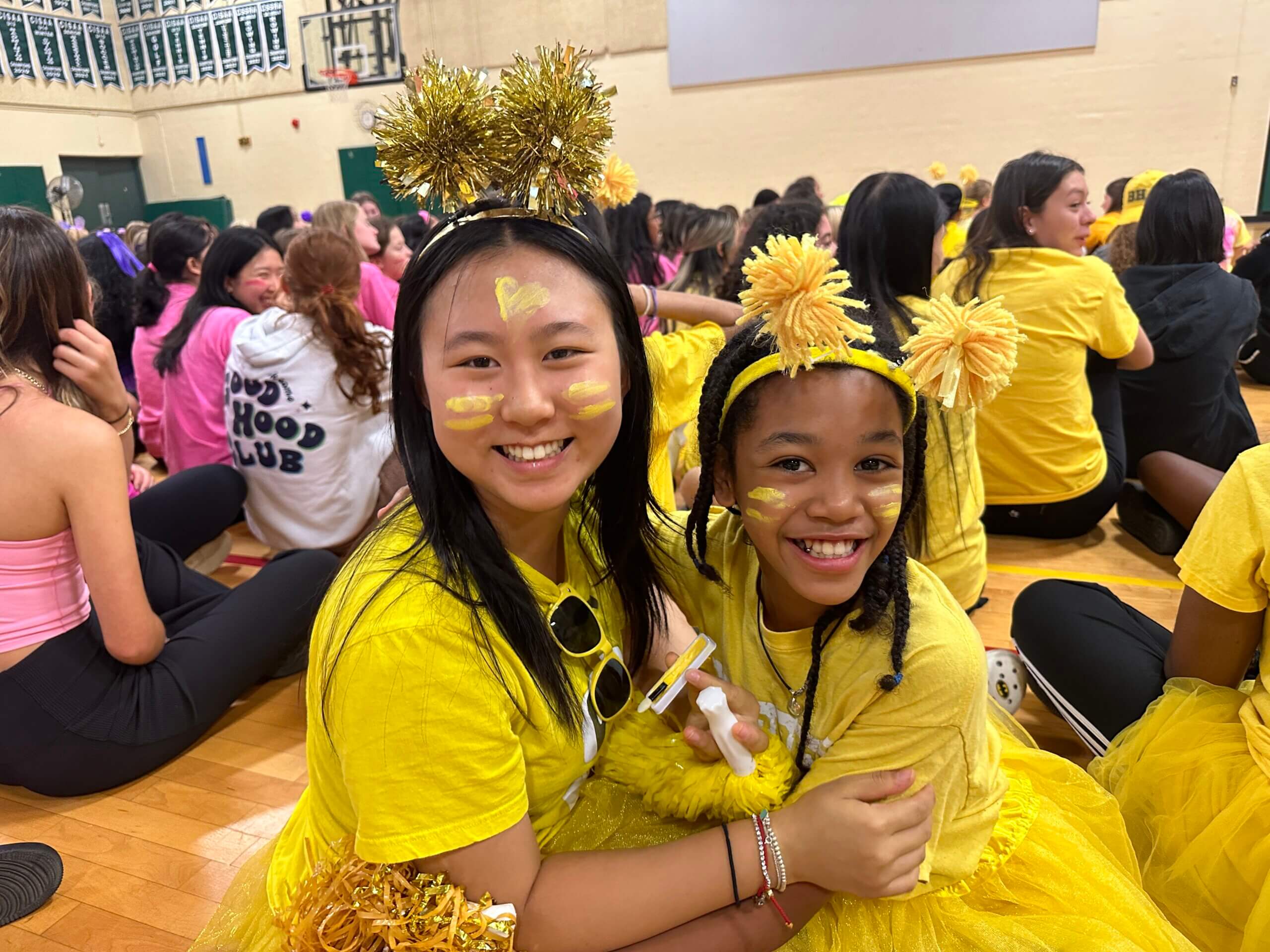 Two girls dressed in yellow outfits, tutus, and pom-pom headbands smile and hug each other at a school event in a gym. Other students, many also wearing colorful clothes, are seated on the floor in the background.
