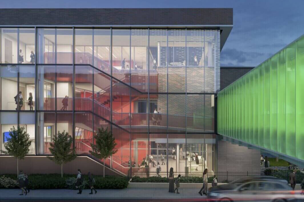 Modern building with large glass windows showing red stairs and people inside; a glowing green walkway connects the building to another structure; several people walk outside near trees and a passing car.