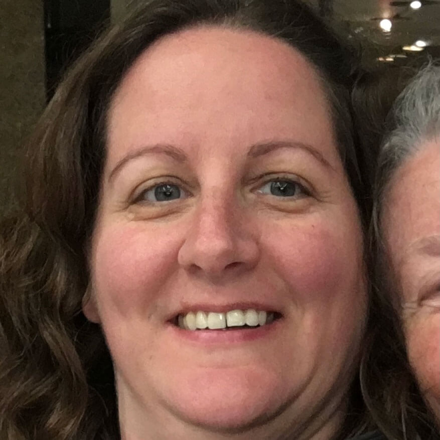 A woman with wavy brown hair smiles at the camera in a close-up photo. Only part of another person's face is visible on the right edge of the image.