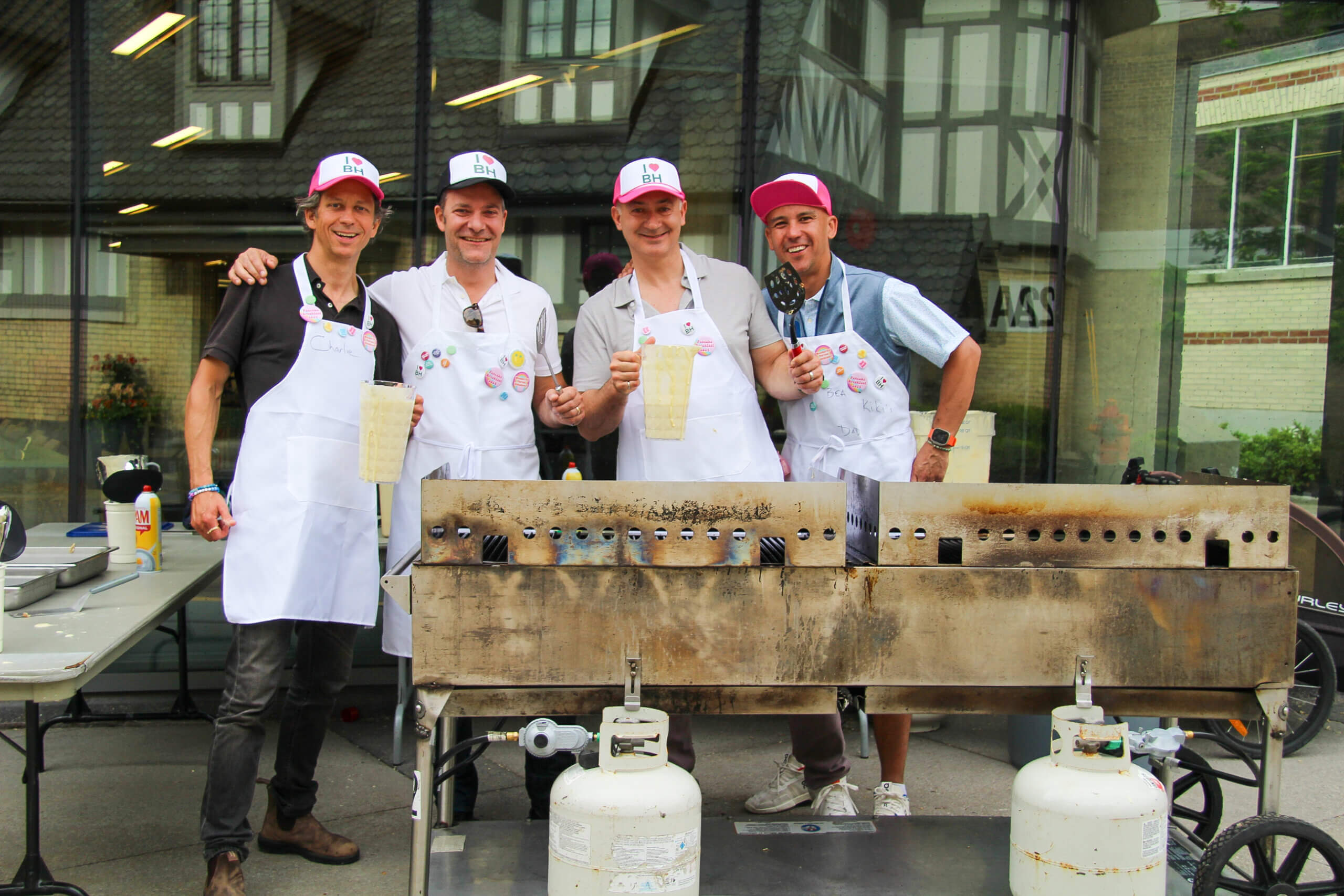 Four men wearing aprons and colorful hats smile while standing behind a large outdoor grill with propane tanks, holding pitchers of batter in front of a building with large windows.