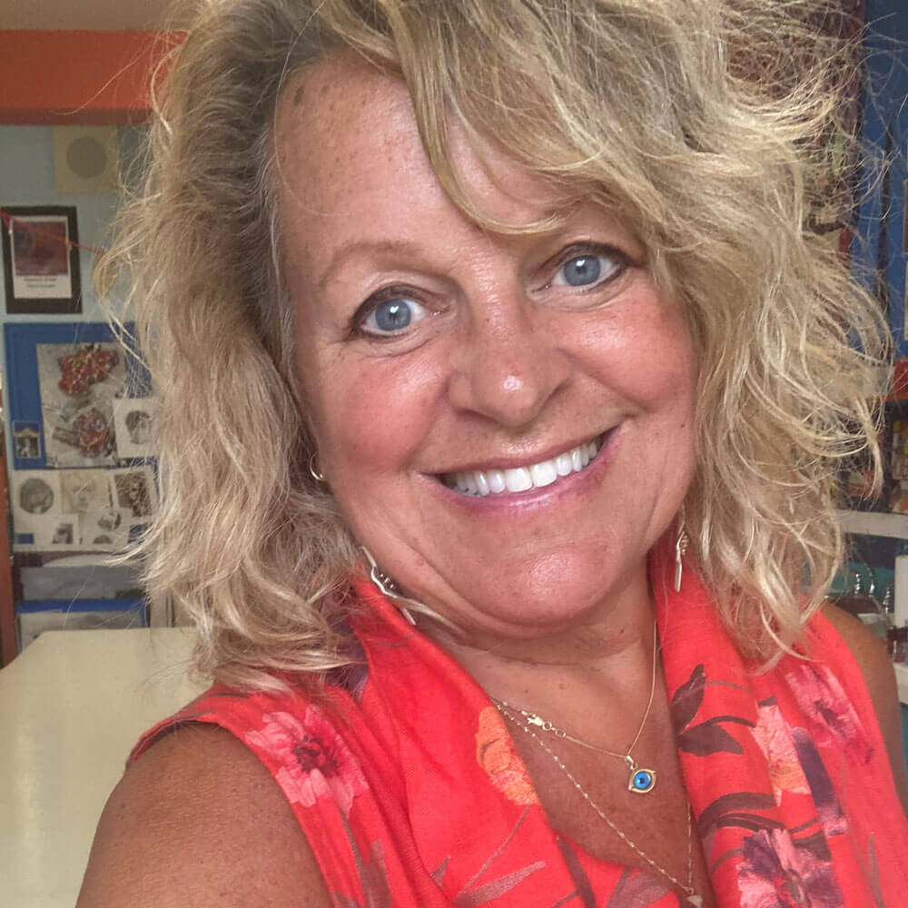 A smiling woman with wavy blonde hair wearing a bright orange floral top and a necklace poses indoors in a colorful room with artwork and photos on the wall behind her.