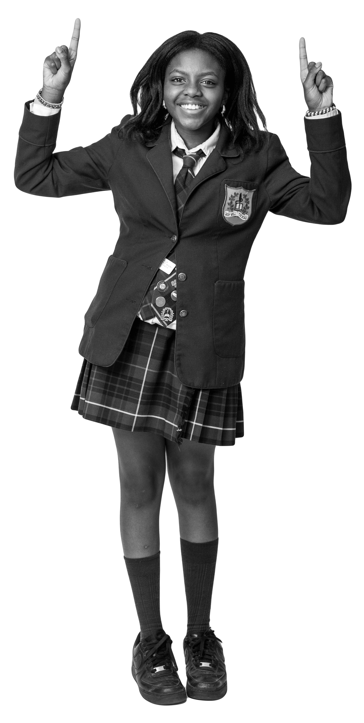 A smiling girl in a school uniform with a blazer and plaid skirt stands pointing both index fingers upward.