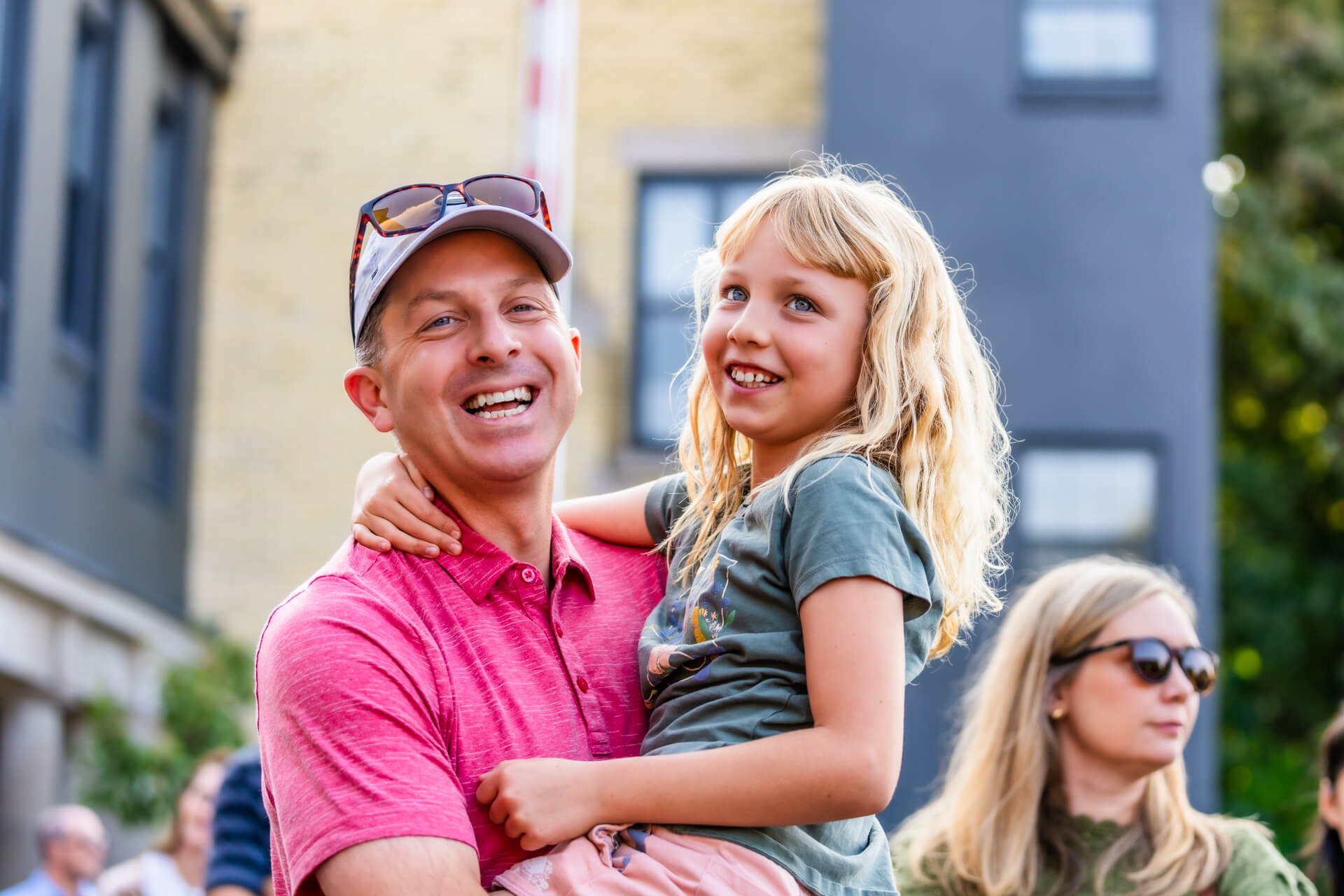 A smiling man in a pink shirt and cap holds a laughing young girl with blond hair at an outdoor event, with people and buildings in the background.