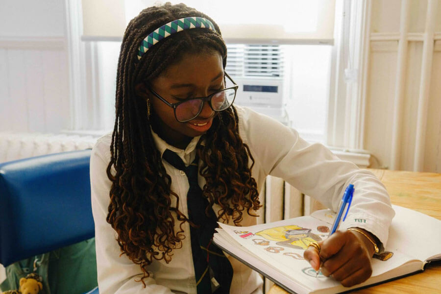 A young girl with glasses and long braids smiles as she draws in a sketchbook with a blue pen, sitting at a table in a brightly lit room.