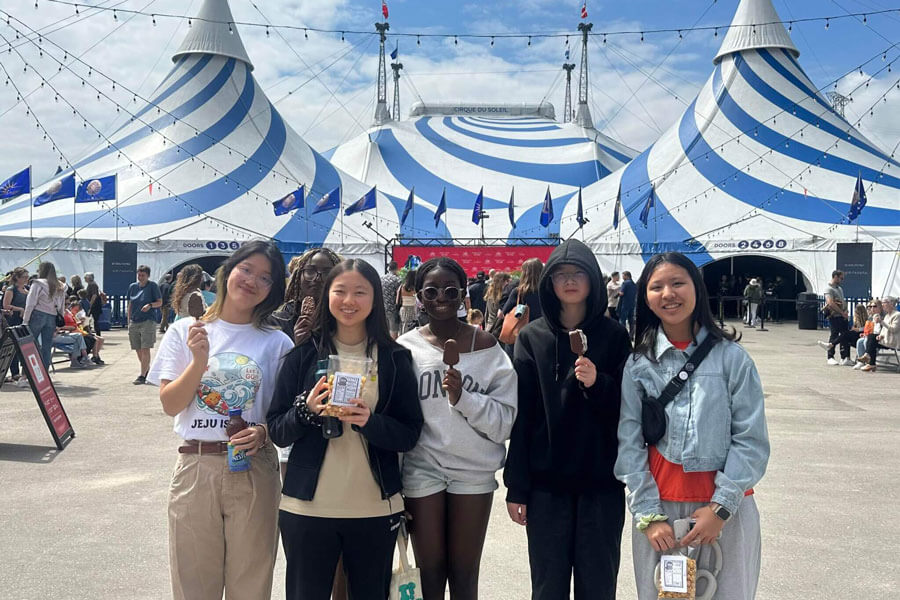 Five young people stand smiling and holding ice cream in front of a large blue and white striped circus tent on a sunny day, with a crowd gathered around the entrance.