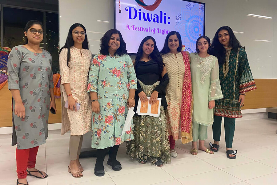 Seven women stand together indoors, smiling in front of a screen displaying “Diwali: A Festival of Lights.” They wear colorful traditional South Asian attire, celebrating the festival.