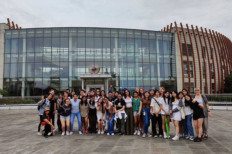 A large group of young people poses and smiles in front of a modern glass building with a curved wooden structure on the right side, under a cloudy sky.