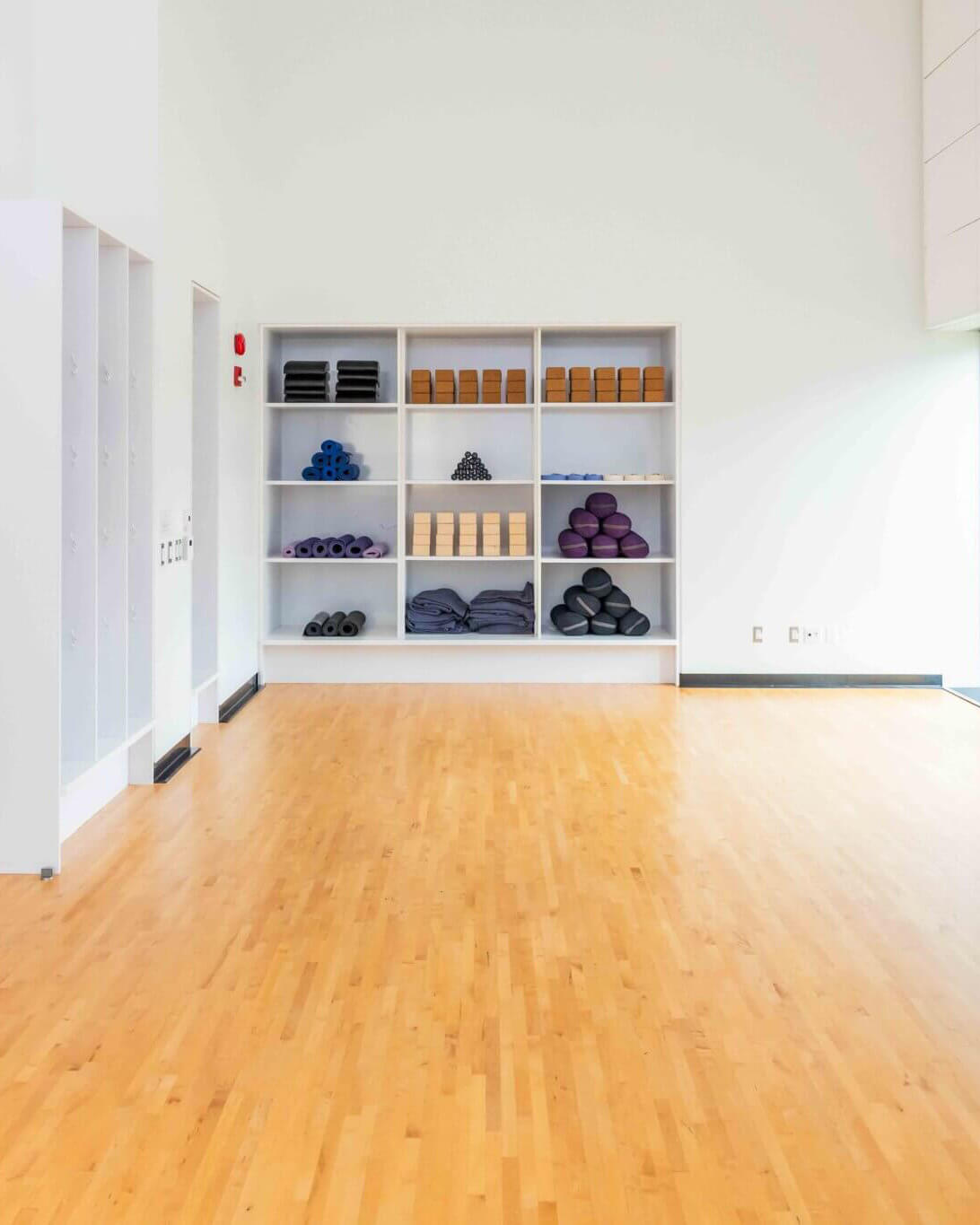 A spacious, bright room with a wooden floor and white walls, featuring shelves neatly stacked with yoga mats, blocks, and blankets. The room is empty and well-lit by natural light.
