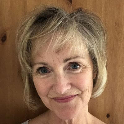 A smiling older woman with short, light blonde hair stands in front of a wooden background. She has blue eyes and is looking directly at the camera.
