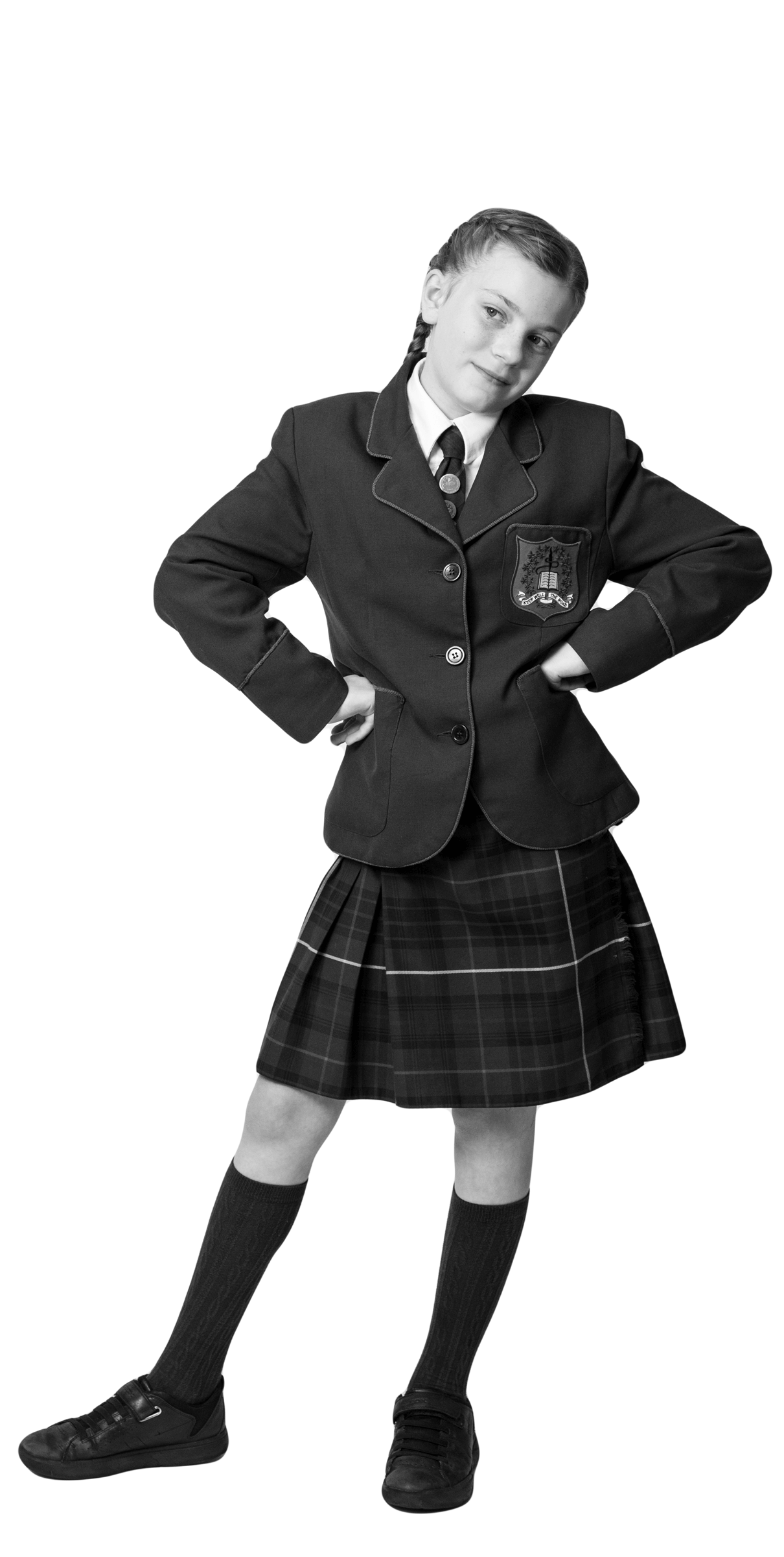 A child in a school uniform with a blazer, tie, pleated skirt, knee-high socks, and dress shoes stands with hands on hips, smiling confidently. The image is in black and white.