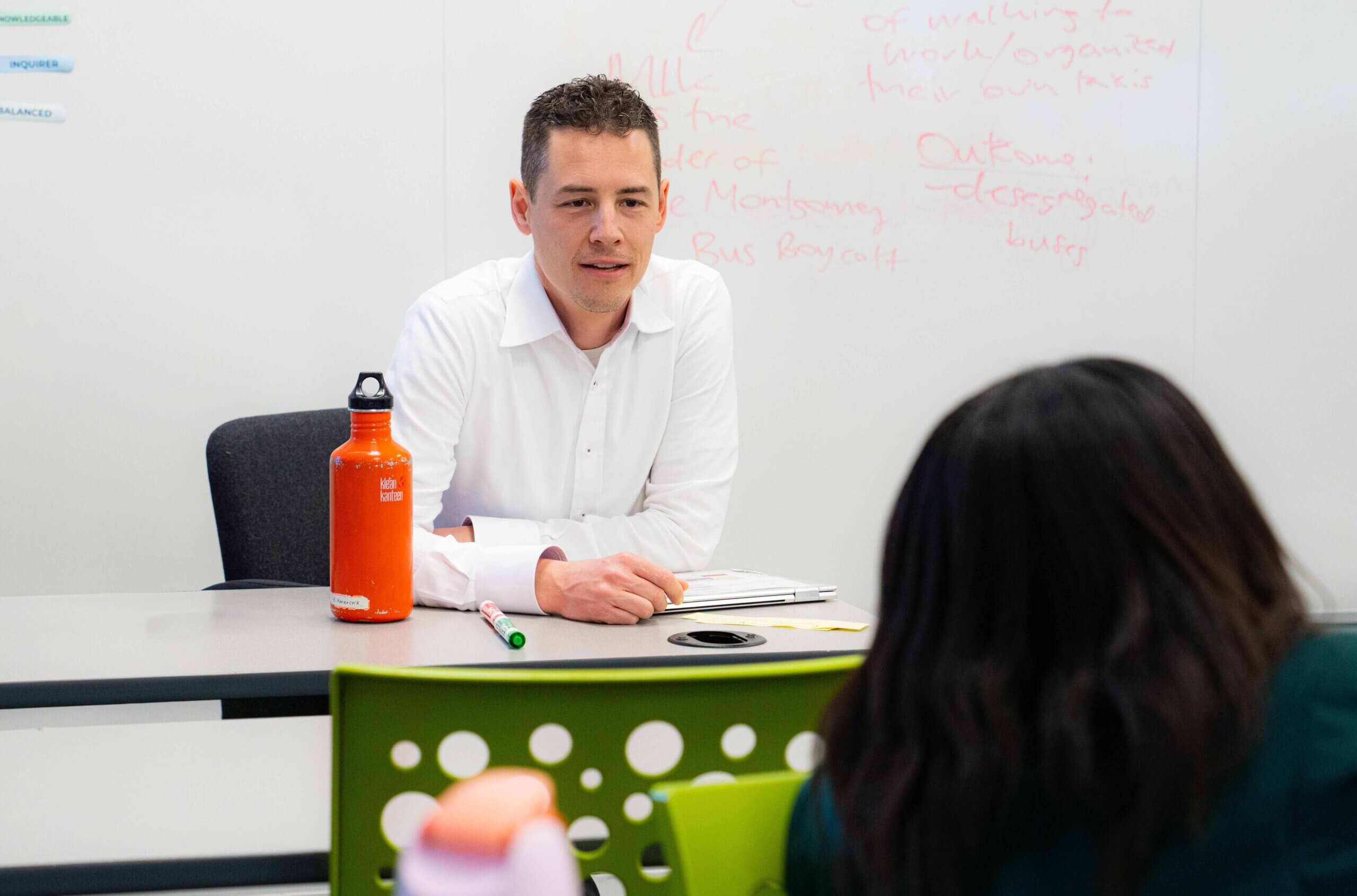 A man in a white shirt sits at a desk with a bright orange water bottle, facing a person with long dark hair. There is writing on a whiteboard behind him. They appear to be in a classroom or meeting setting.