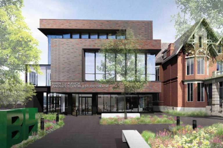 Modern brick and glass building labeled "Karen Hjurjevich Innovation Centre & Studio Theatre" beside a traditional red-brick house, set amid landscaped gardens and trees on a sunny day.