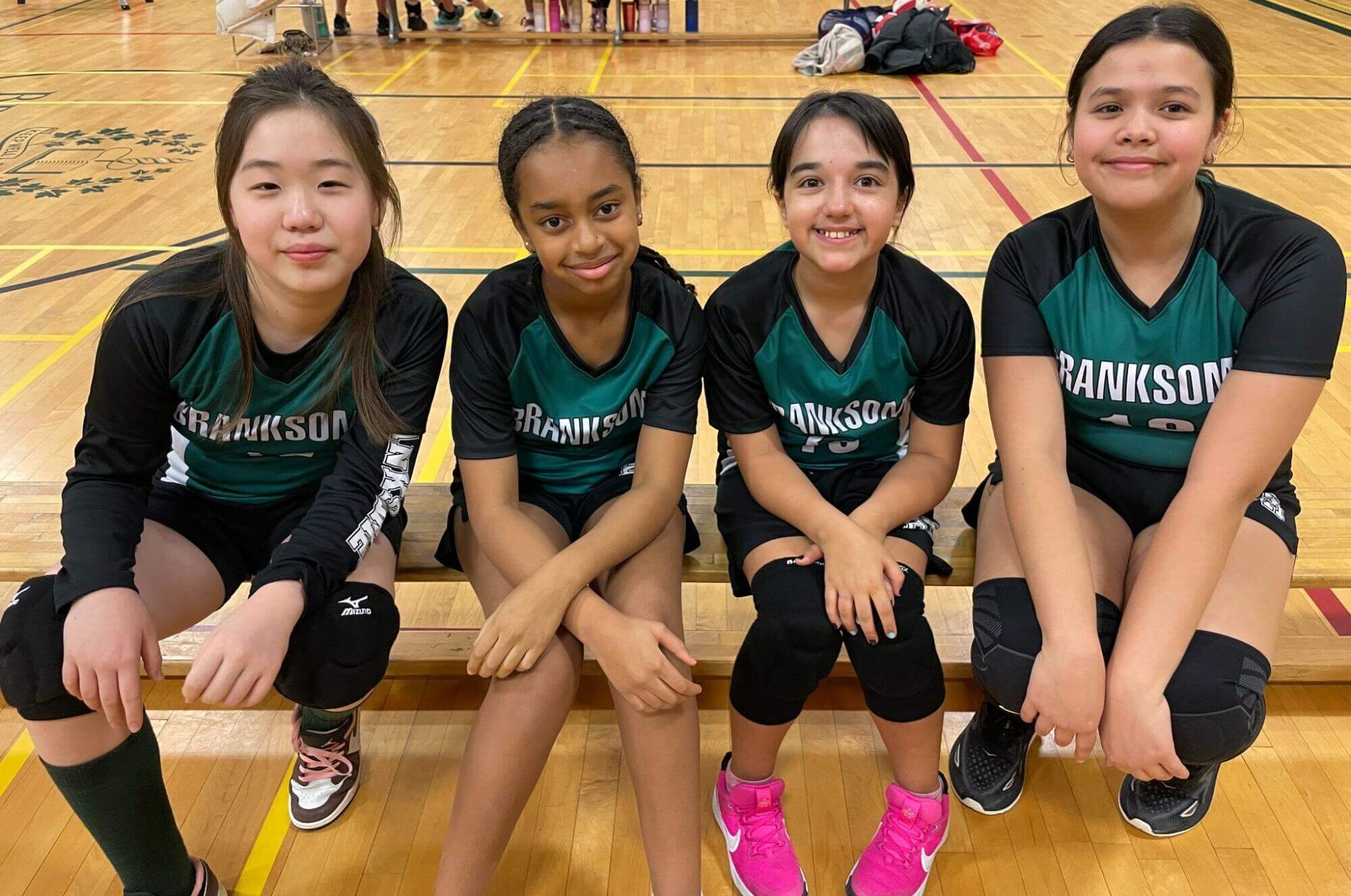 Four girls in matching green and black sports uniforms sit on a bench in a gym, smiling at the camera. Volleyball knee pads and shoes are visible, and other players and water bottles are in the background.