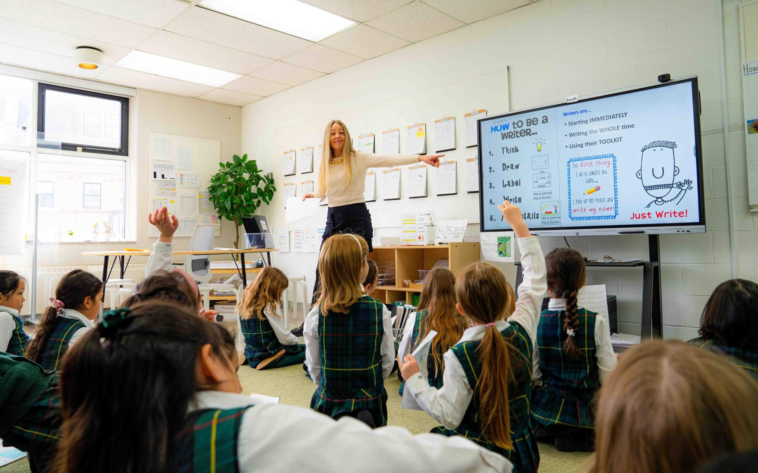 A teacher stands at the front of a classroom, smiling and pointing to a board, while several students in plaid uniforms sit on the floor with their hands raised, eager to participate.