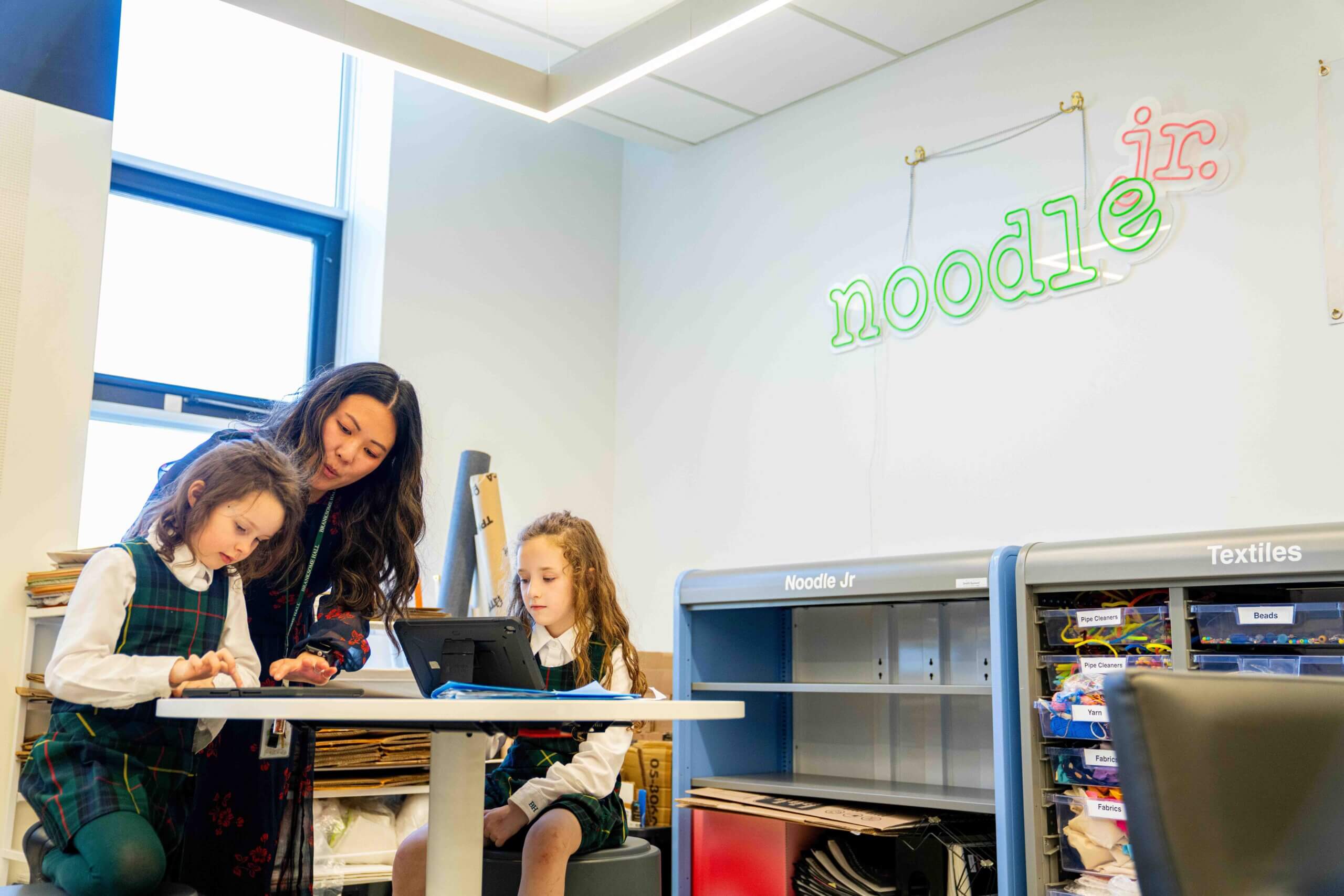 A teacher helps two young girls in school uniforms with a project at a table in a classroom labeled “noodle jr.” on the wall. One girl is seated, while the other stands next to the teacher looking at a tablet.