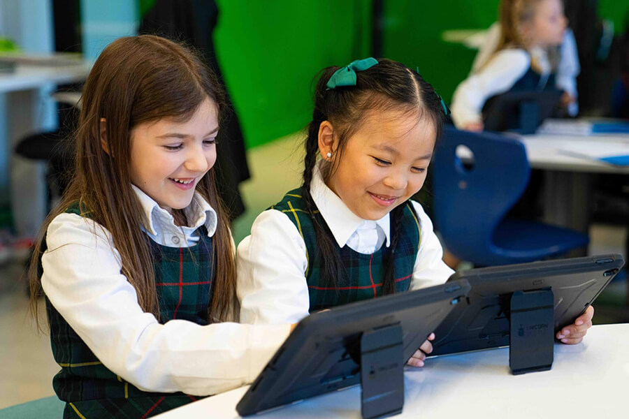 Two young girls in school uniforms sit side by side at a table, smiling and using tablet devices in a brightly lit classroom. Other students and desks are visible in the background.