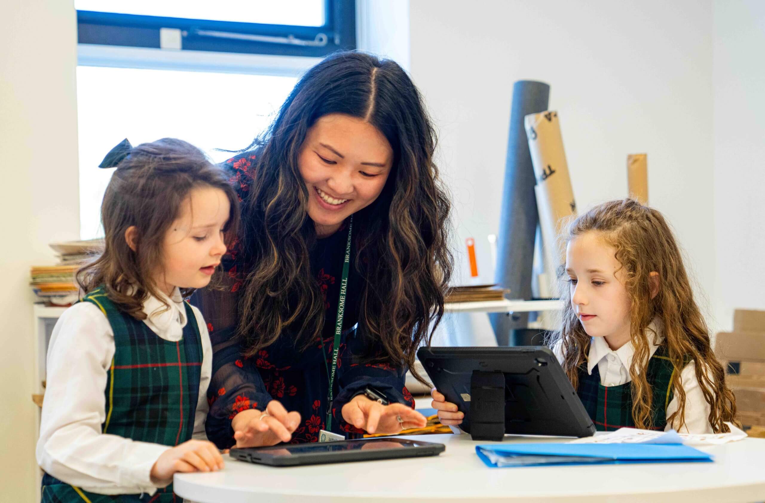 A teacher smiles while helping two young girls in plaid uniforms use tablets at a round table in a classroom. The girls are focused on their screens, and school supplies are visible in the background.