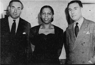 Three people pose for a black-and-white photo. A woman stands in the center, wearing a dark dress, flanked by two men in suits and ties on either side. All three look directly at the camera.