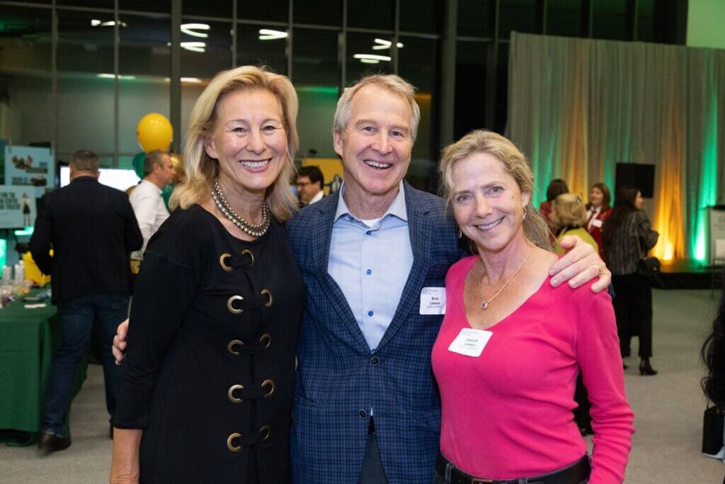 Three smiling adults, two women and one man, stand close together at an indoor event. People and tables are visible in the background, with green and yellow lighting creating a festive atmosphere.