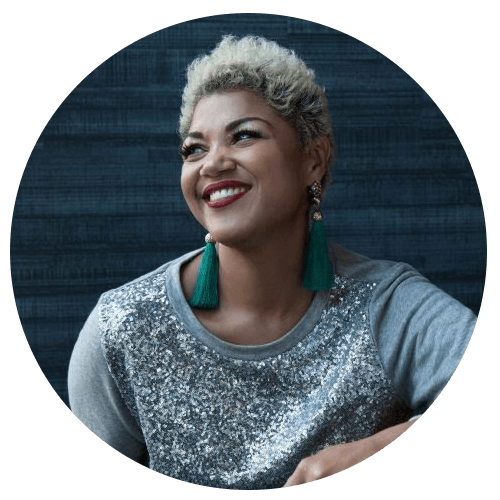 A smiling woman with short, curly blond hair wears a sparkly silver top and large green tassel earrings, sitting against a dark blue textured background.