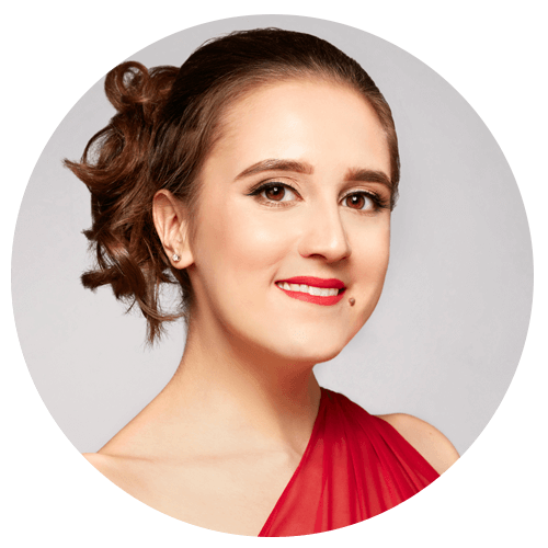 A woman with brown hair styled in an updo, wearing a red, one-shoulder dress and red lipstick, smiling at the camera against a light gray background.